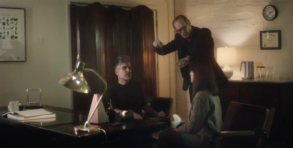 Hayden and Feist sit in an office, while Steve Buscemi stands between them holding a ring on a necklace as if to hypnotize them
